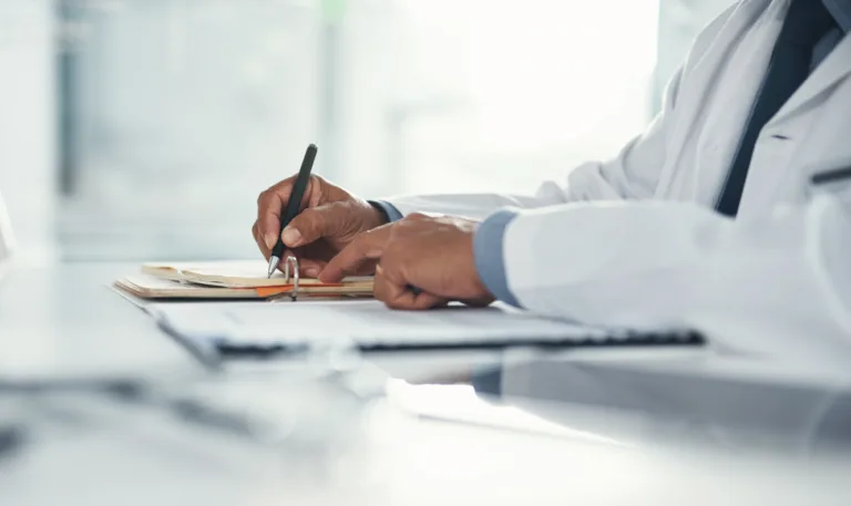 Doctor hands writing on paper or document at a desk in the hospital. Healthcare professional drafting a medical insurance letter, legal paperwork or form. A GP filing a document in a clinic office