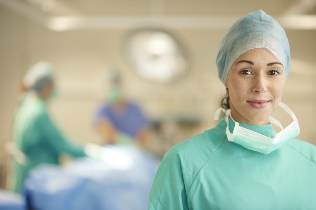 a proud female operating nurse stands away from the operating table and smiles to camera. In the background the anaesthetist and surgeon are mopping up guts after a succesful operation .