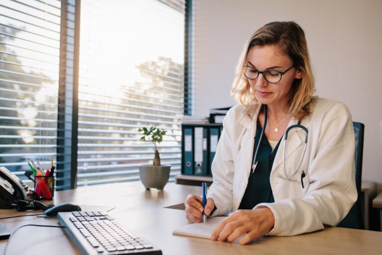 A female doctor with glasses, wearing a white coat and stethoscope, writing in a notebook at her desk in an office with blinds and a computer.