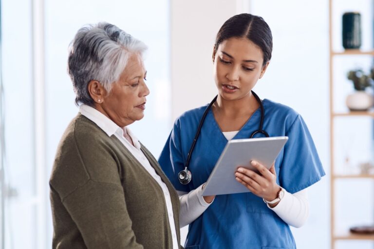 A nurse in blue scrubs shows a digital tablet to an elderly woman in a gray jacket, discussing health information in a clinic setting.