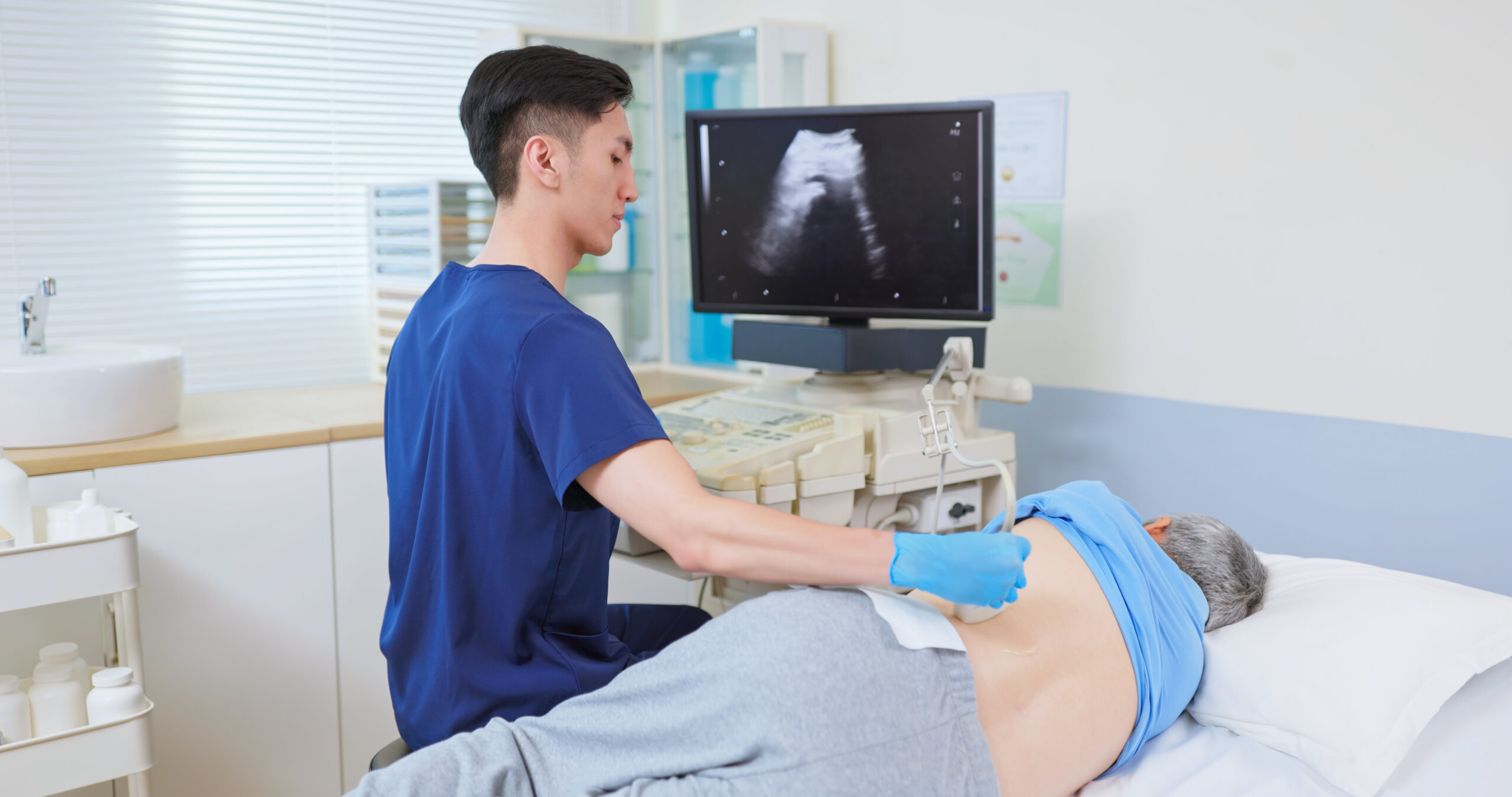 asian old man is in clinic lying side on treatment bed - the young male urologist uses kidney ultrasound to check his health
